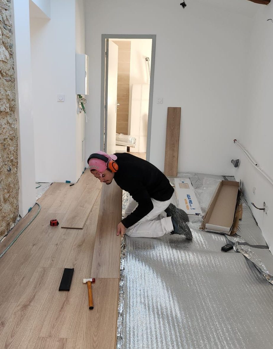 Pose d’un parquet stratifié par un artisan professionnel dans une pièce rénovée avec mur en pierre apparente et poutres en bois, à Marseille